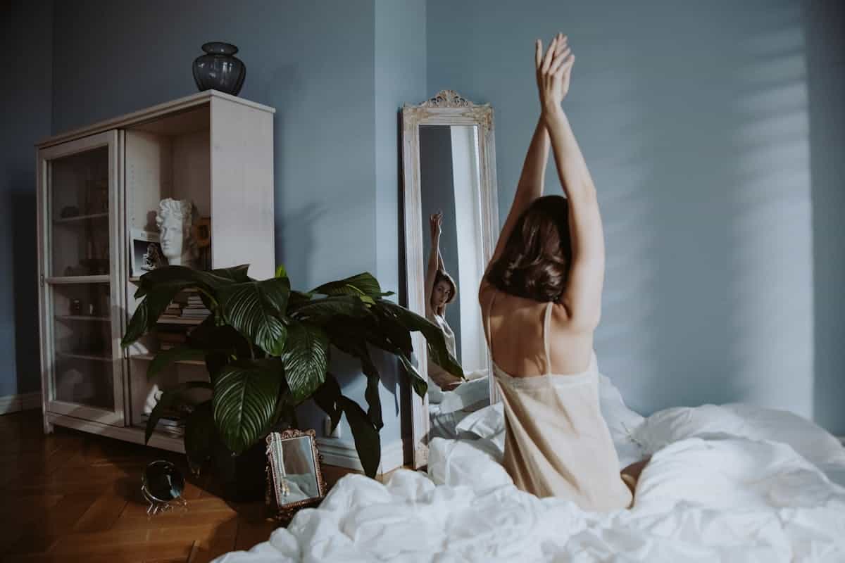 A woman in sleepwear stretches in a calm bedroom setting, reflected in a mirror