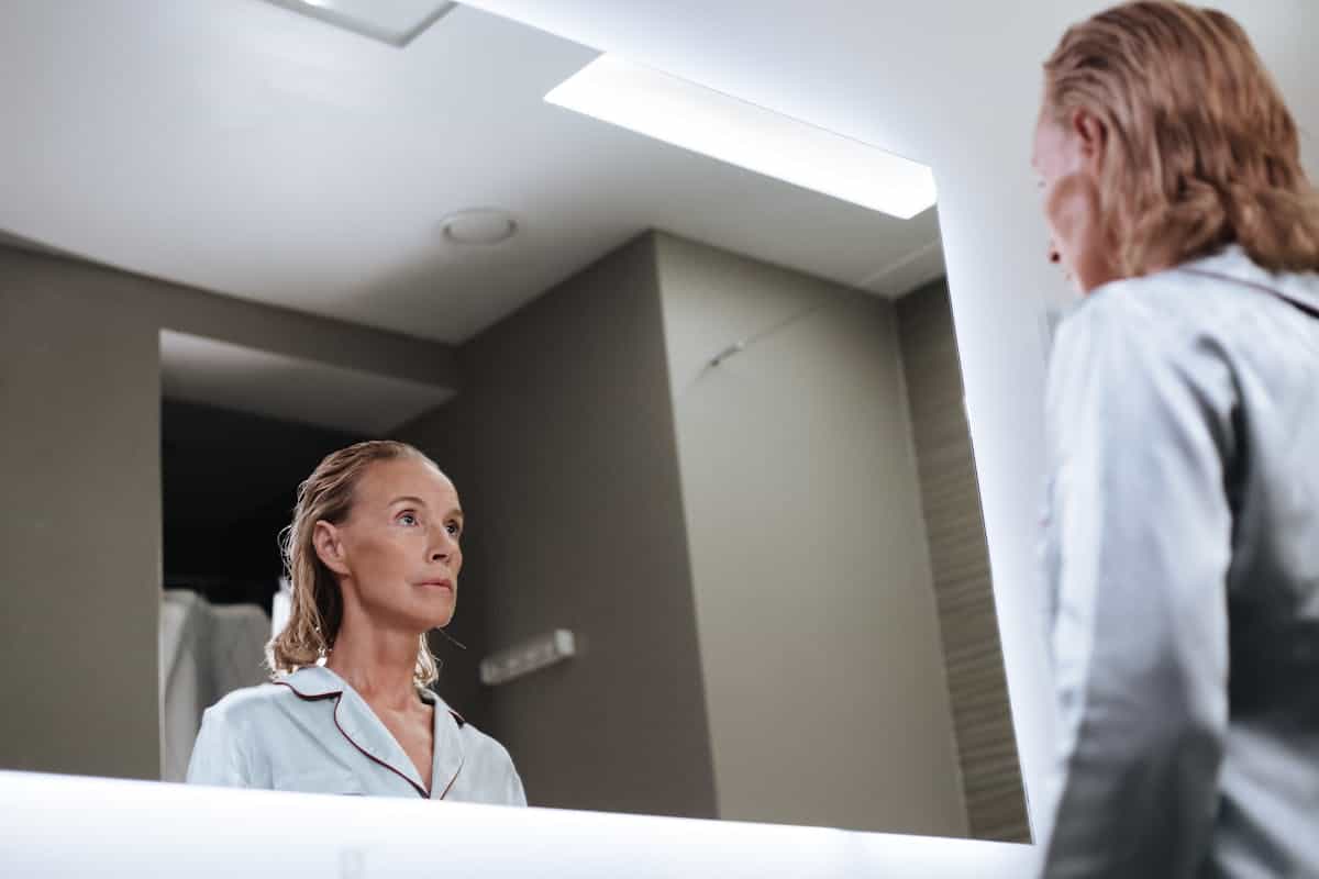 Woman looking introspectively at her bathroom mirror evoking the self-confrontational act of talking to a mirror and its psychological meaning A senior woman looks introspectively at herself in a modern bathroom mirror in quiet indoor lighting evoking the reflective quality of talking to a mirror and what it means