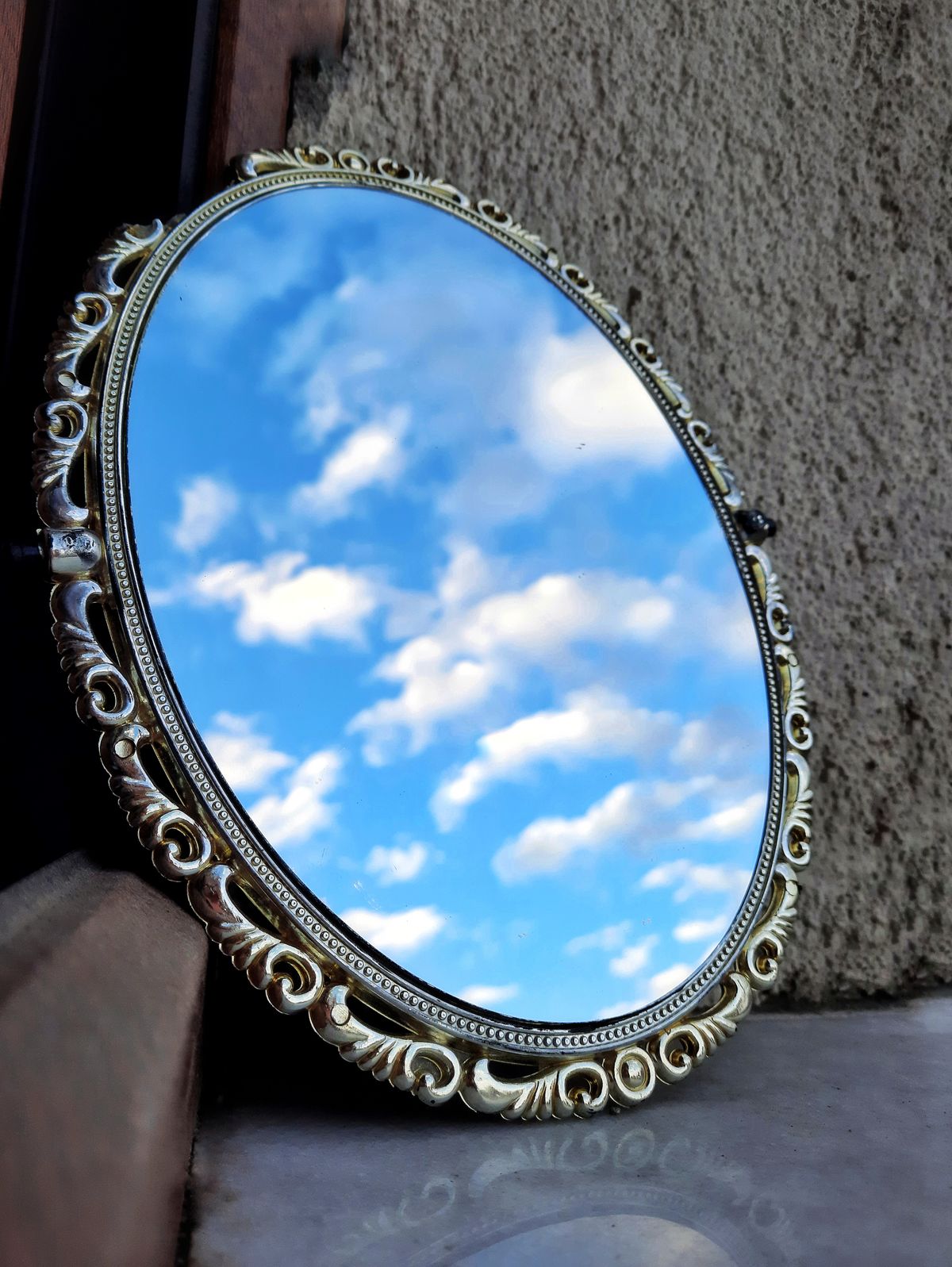 Ornate round mirror reflecting soft blue sky and clouds outdoors