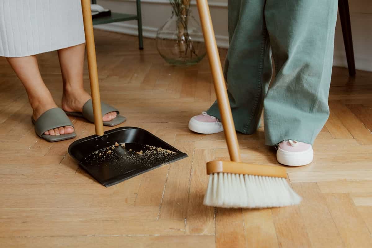 Close-up of people cleaning wooden floor with broom and dustpan carefully collecting broken glass representing the practical and spiritual first steps after a mirror breaks