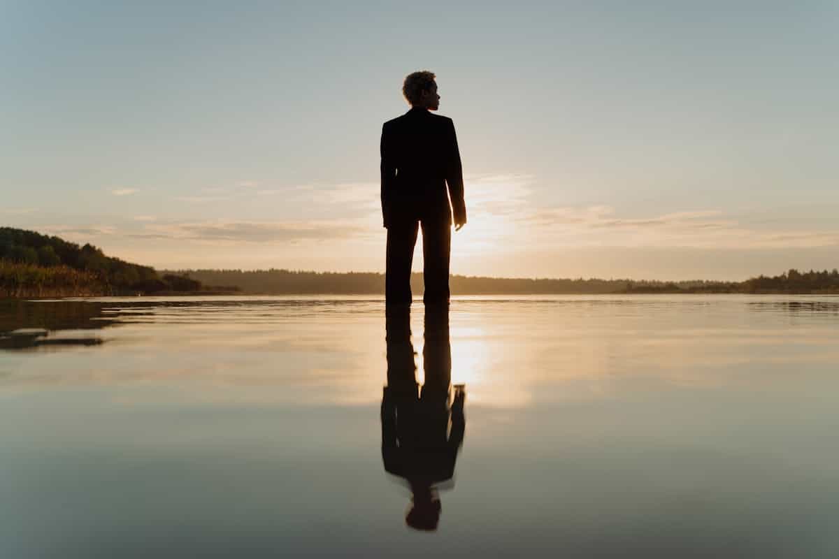 A dramatic silhouette of a figure standing on calm water at sunset reflected in the surface representing the shadow self and unconscious aspects revealed in mirror-in-water dreams