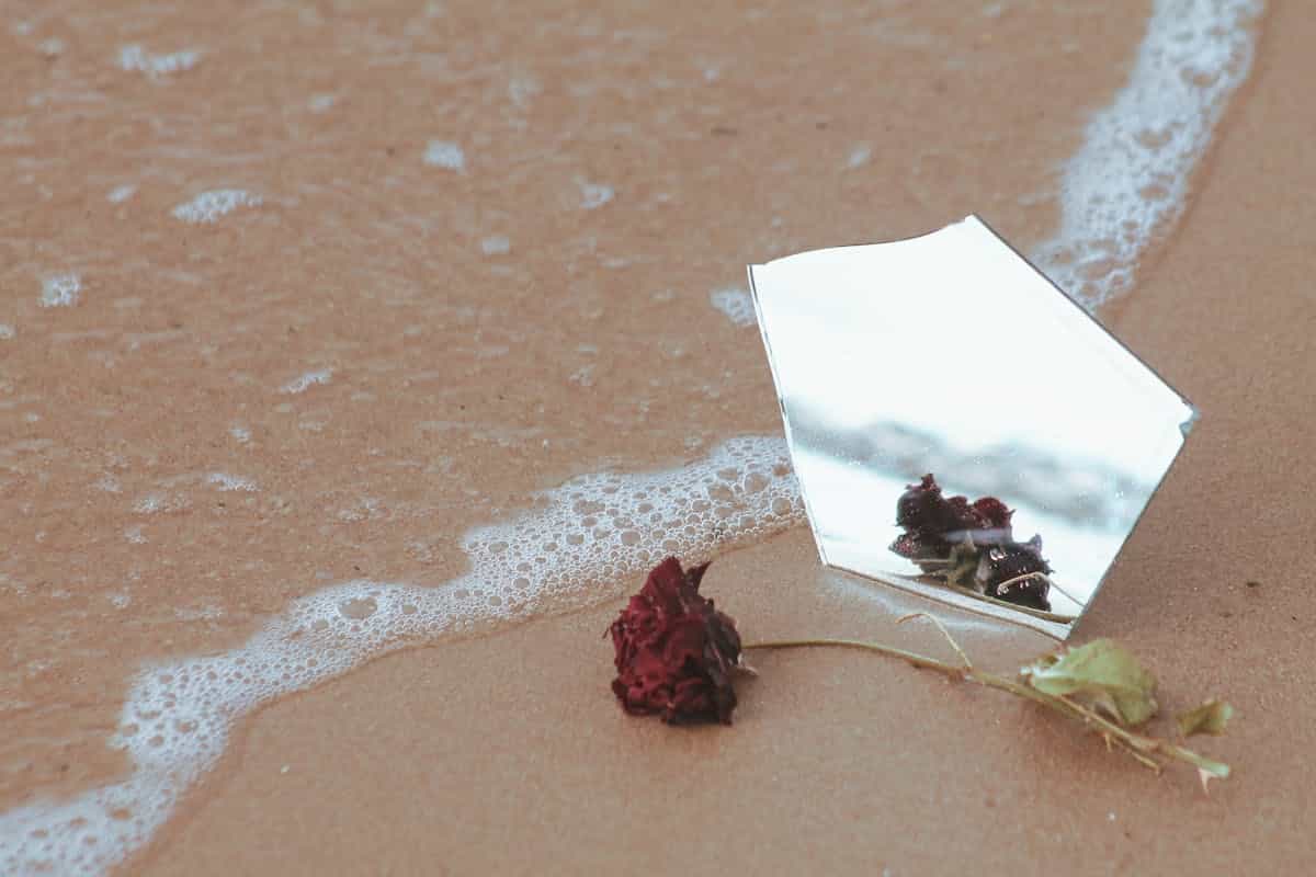A broken mirror reflecting on sand with a red rose on a beach shore representing the complexity and beauty of broken mirror symbolism in art literature and culture