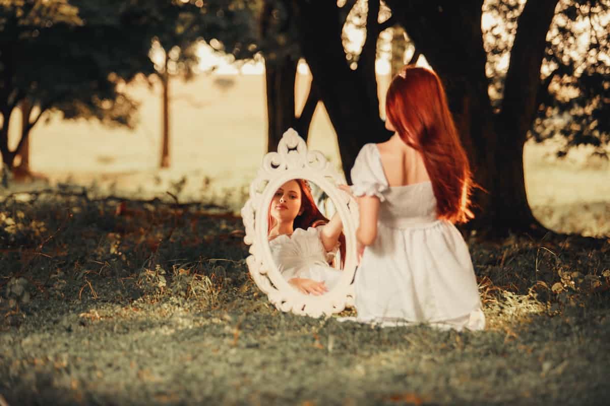 Woman with mirror outdoors in nature evoking the spiritual invitation that recurring mirror dreams extend across traditions A redhead woman sits outdoors with a small hand mirror reflecting the surrounding nature evoking the spiritual dimension of dreams about mirrors and what they ask us to examine