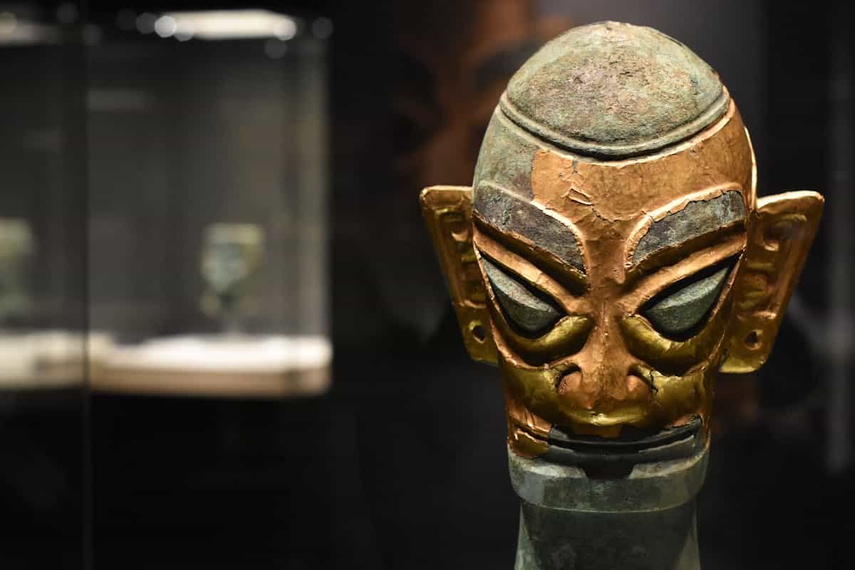 Close-up of an ancient bronze mask in a dimly lit museum display case representing the historical reality of ancient bronze mirrors that informed biblical mirror metaphors