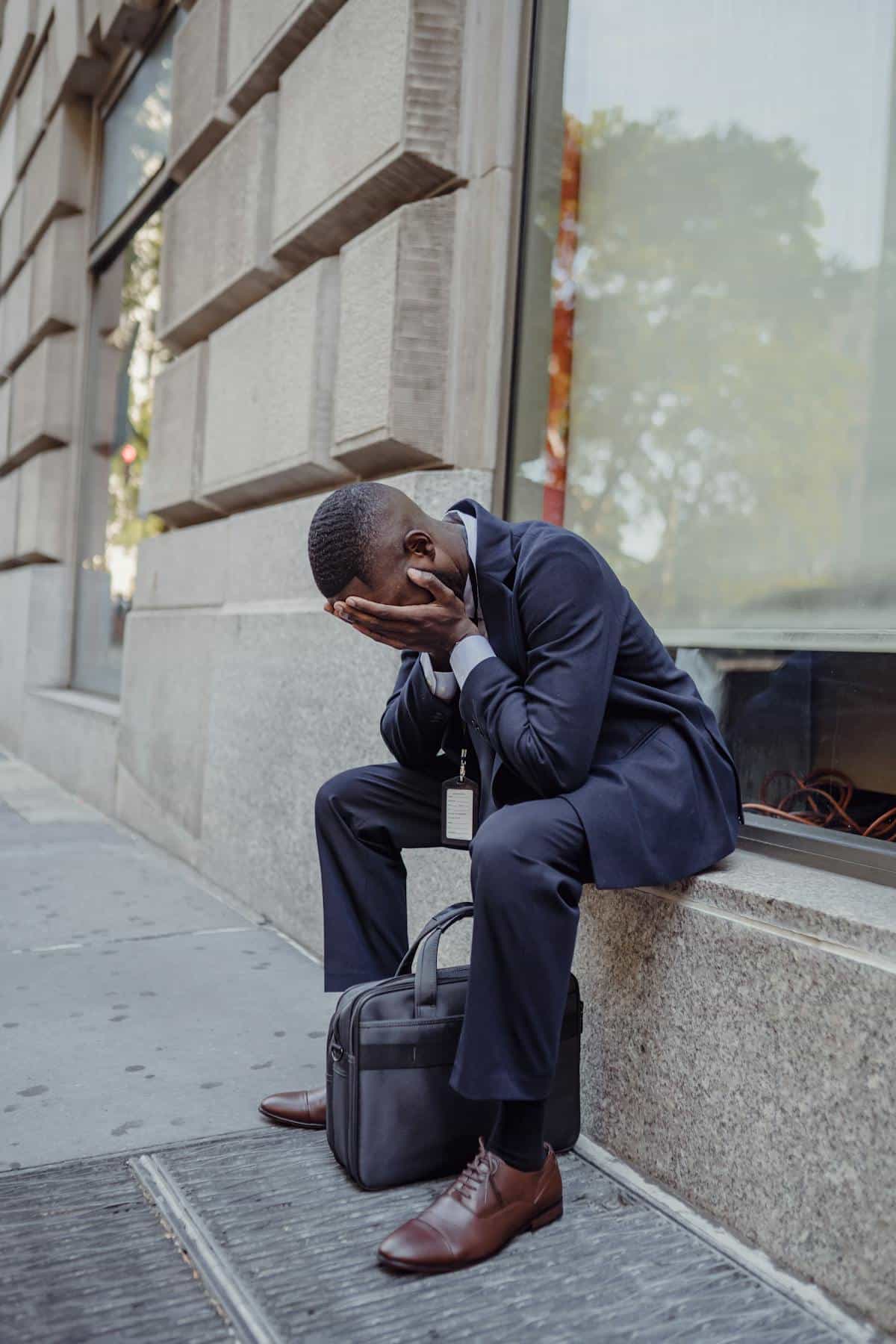 Stressed professional on city sidewalk illustrating work identity strain and dream stress imagery Stressed man in business suit sitting on city sidewalk with face in hands reflecting burnout and work anxiety themes