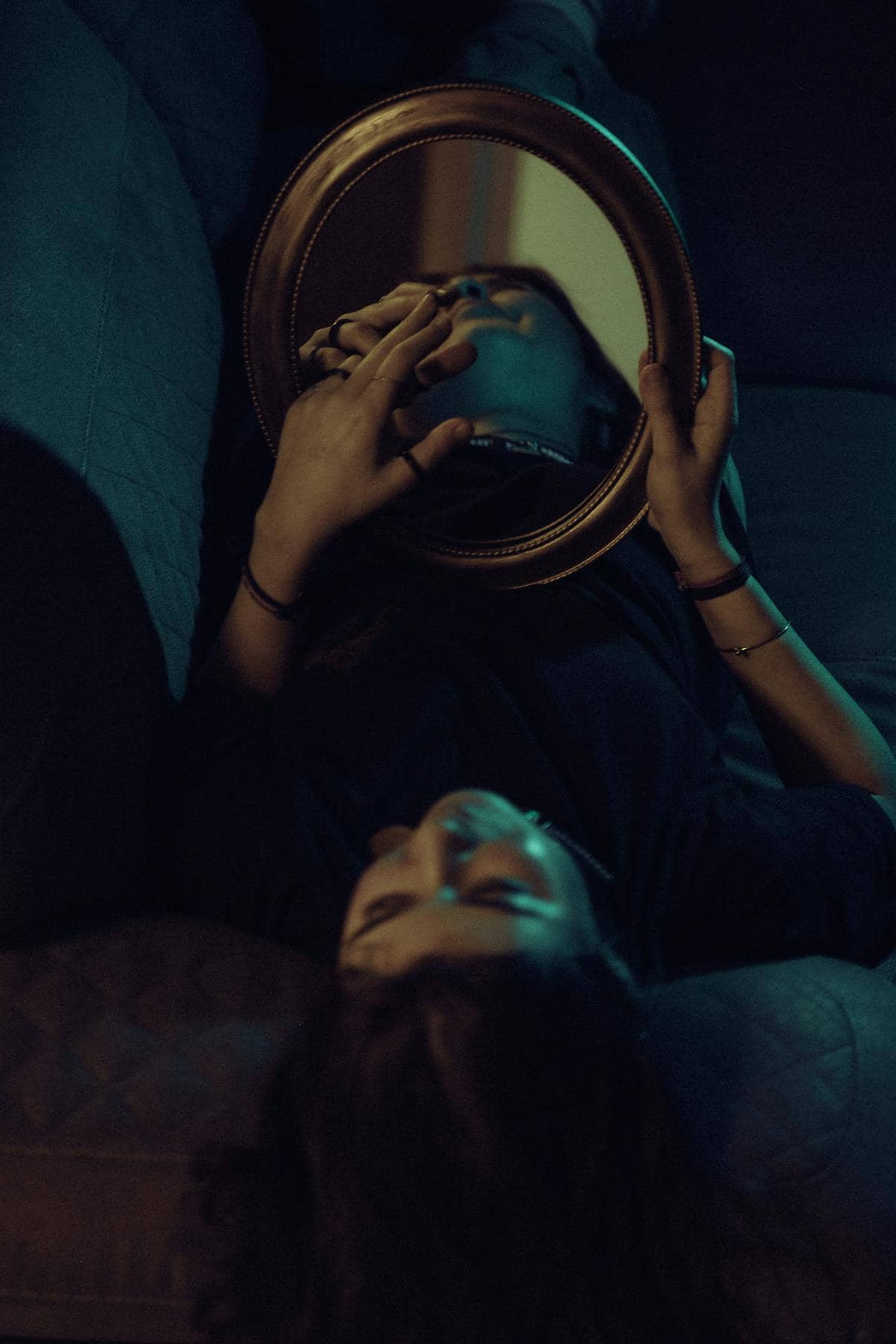 Moody interior scene with a reclining figure holding an oval handheld mirror reflecting dim window light