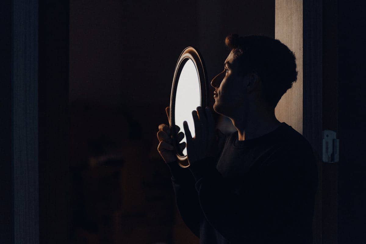 Silhouetted figure holding a mirror at a dark doorway edge suggesting threshold symbolism for mirror meditation closure