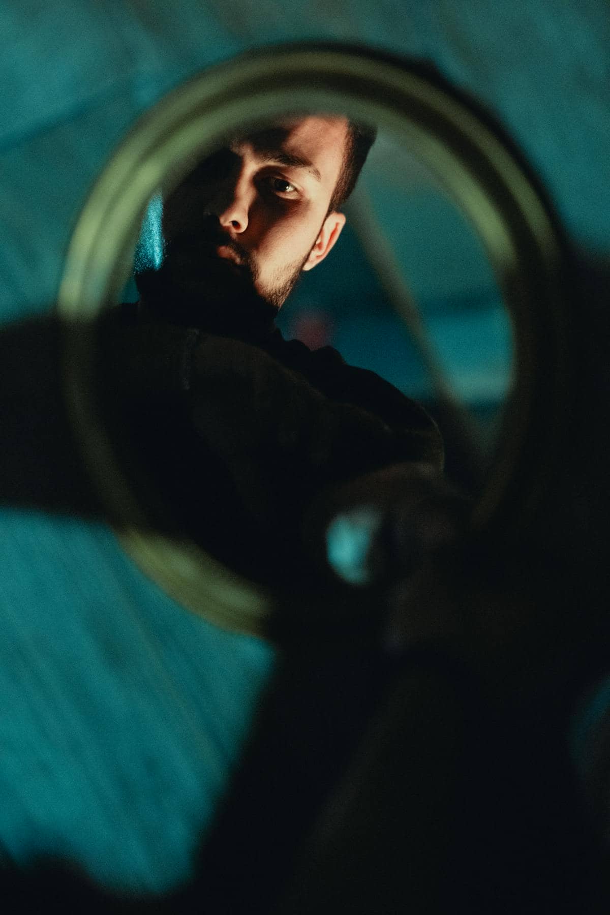 High-angle dramatic reflection study of a seated figure confronting mirror glass in teal cast lighting
