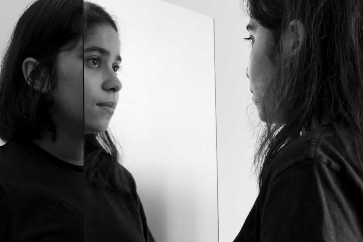 Black and white portrait of a young woman gazing at her own reflection in a mirror indoors representing the psychological theme of self-perception, identity recognition, and honest self-confrontation central to mirror dream interpretation