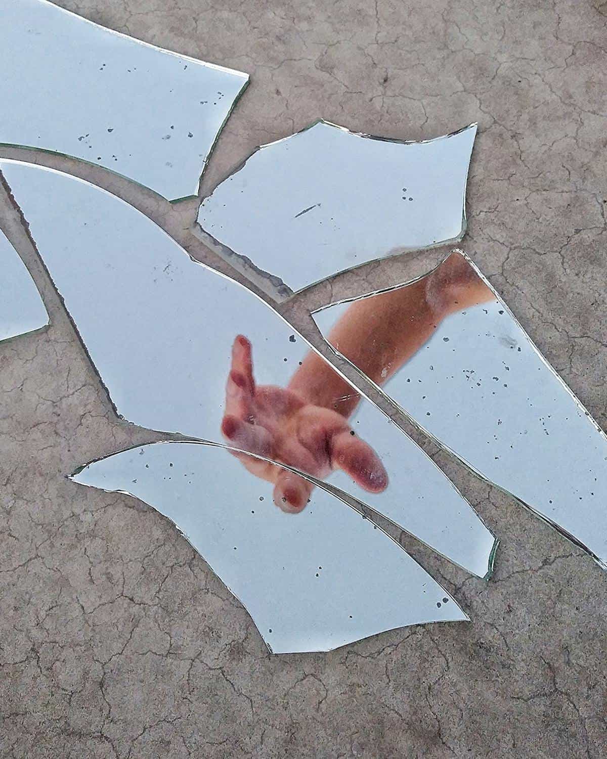 Hand reflections scattered across irregular mirror shards on muted gray tabletop