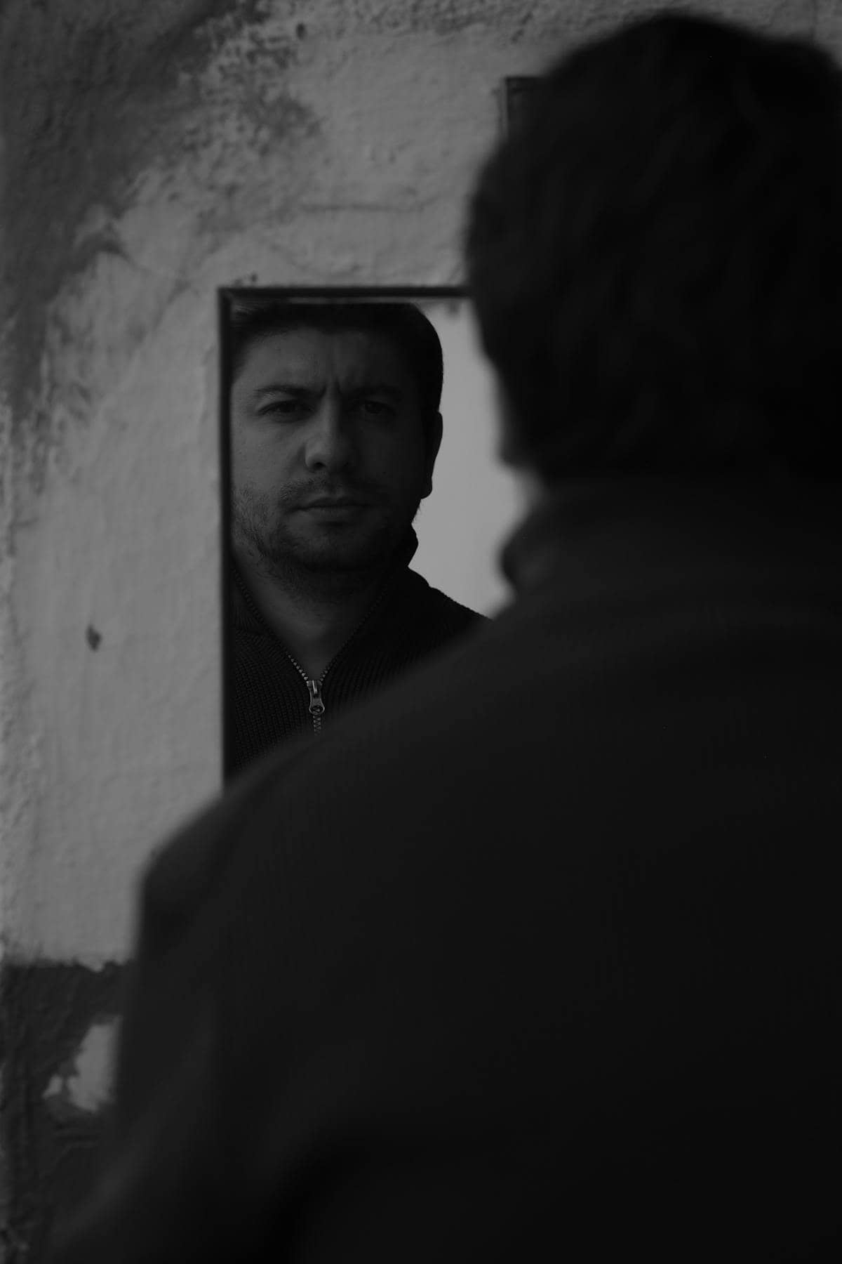 Monochrome mirrored portrait showing a masculine figure inspecting reflection with subdued studio lighting contrasts