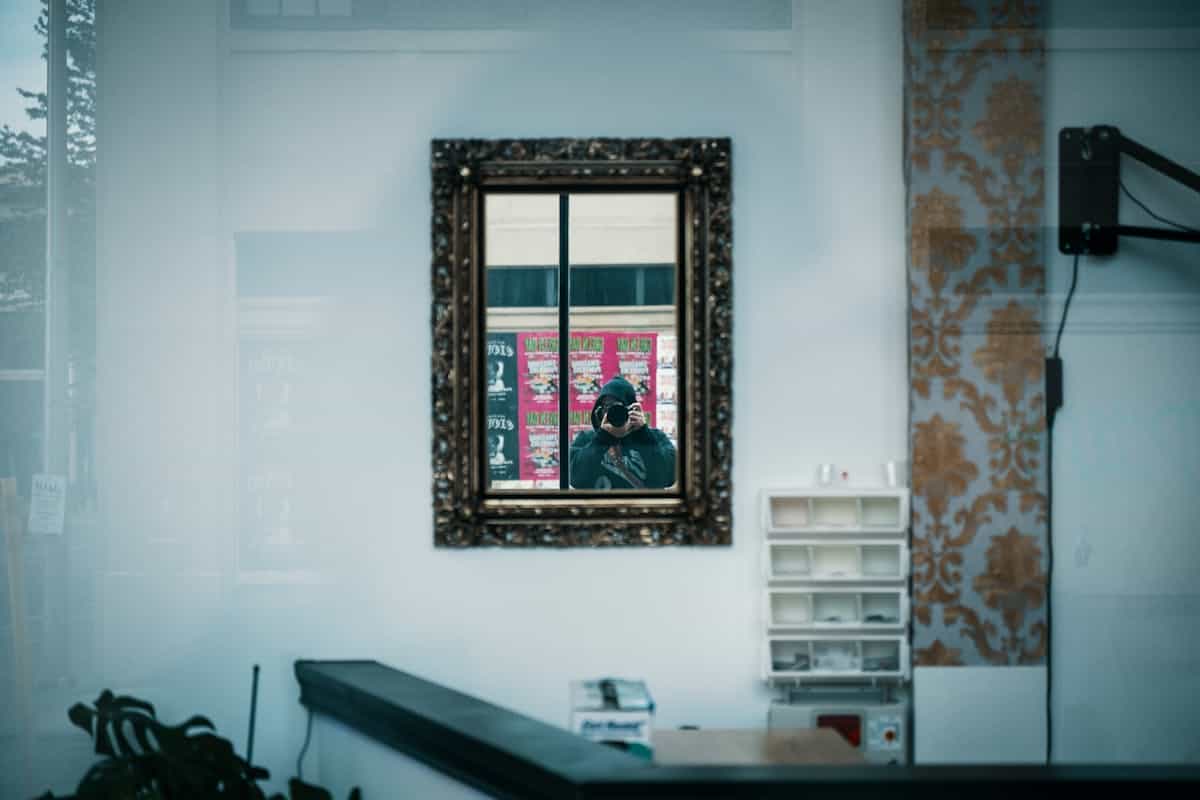 Person in dark knit sweater seen from side gazing into wall mirror in dim interior moody self reflection scene