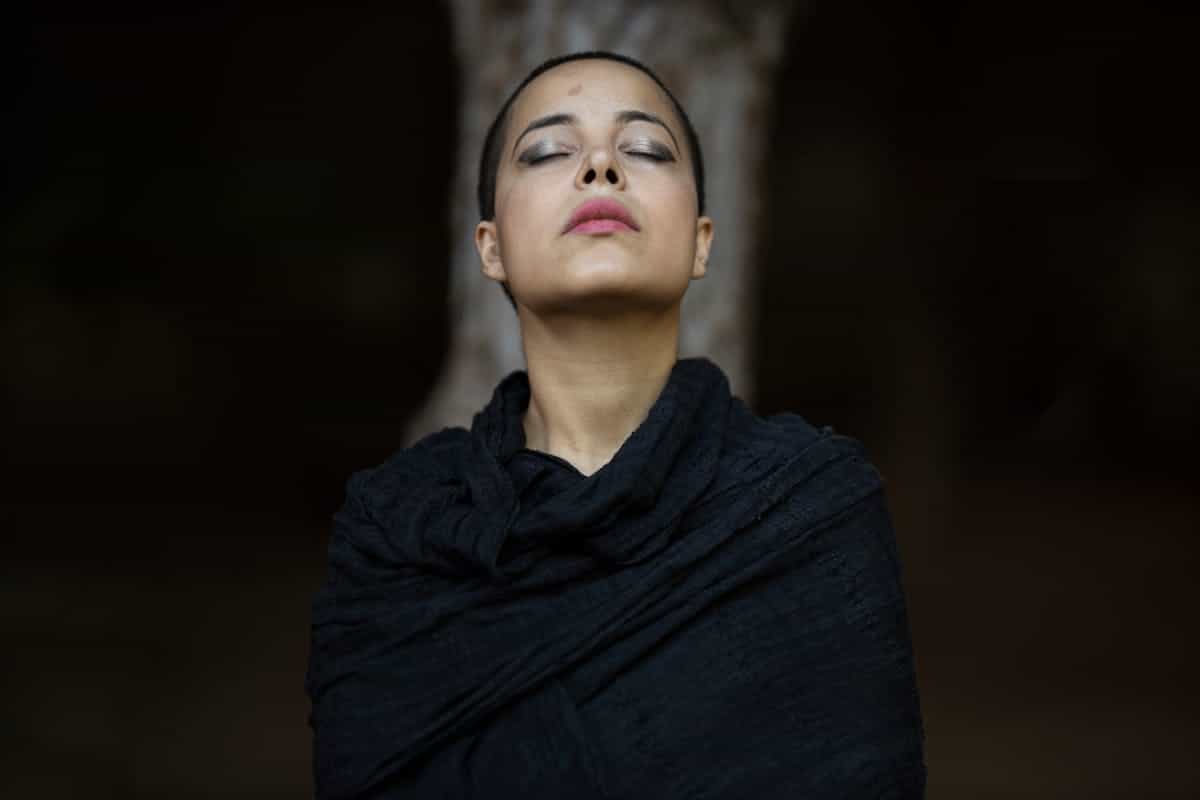 A serene portrait of a woman with eyes closed, wrapped in a shawl against a blurred background, evoking the state of deep receptive stillness required for effective black mirror scrying practice