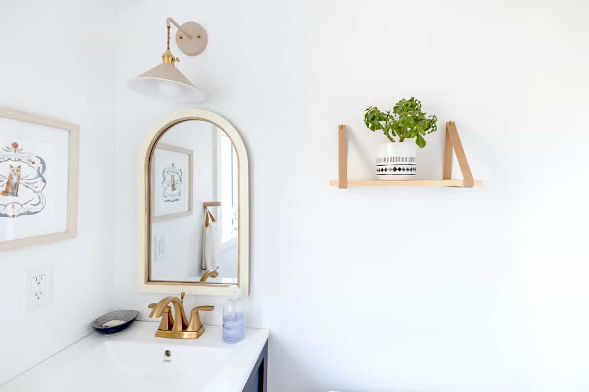 Gold-framed mirror in a bright bathroom illustrating arched mirror frame styles Bright bathroom interior with a gold-framed mirror and a decorative shelf, illustrating arched mirror frame styles
