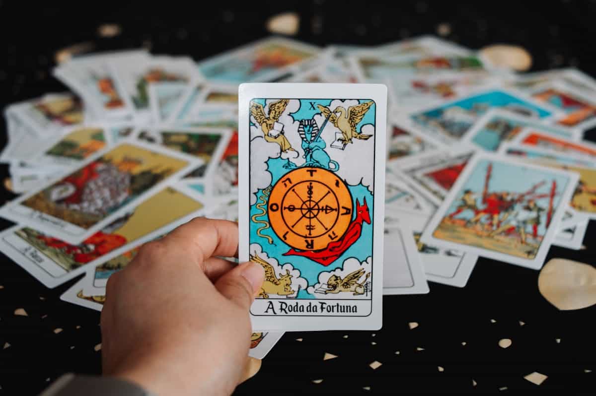 A hand holding a Wheel of Fortune tarot card surrounded by scattered tarot cards on a dark textured surface