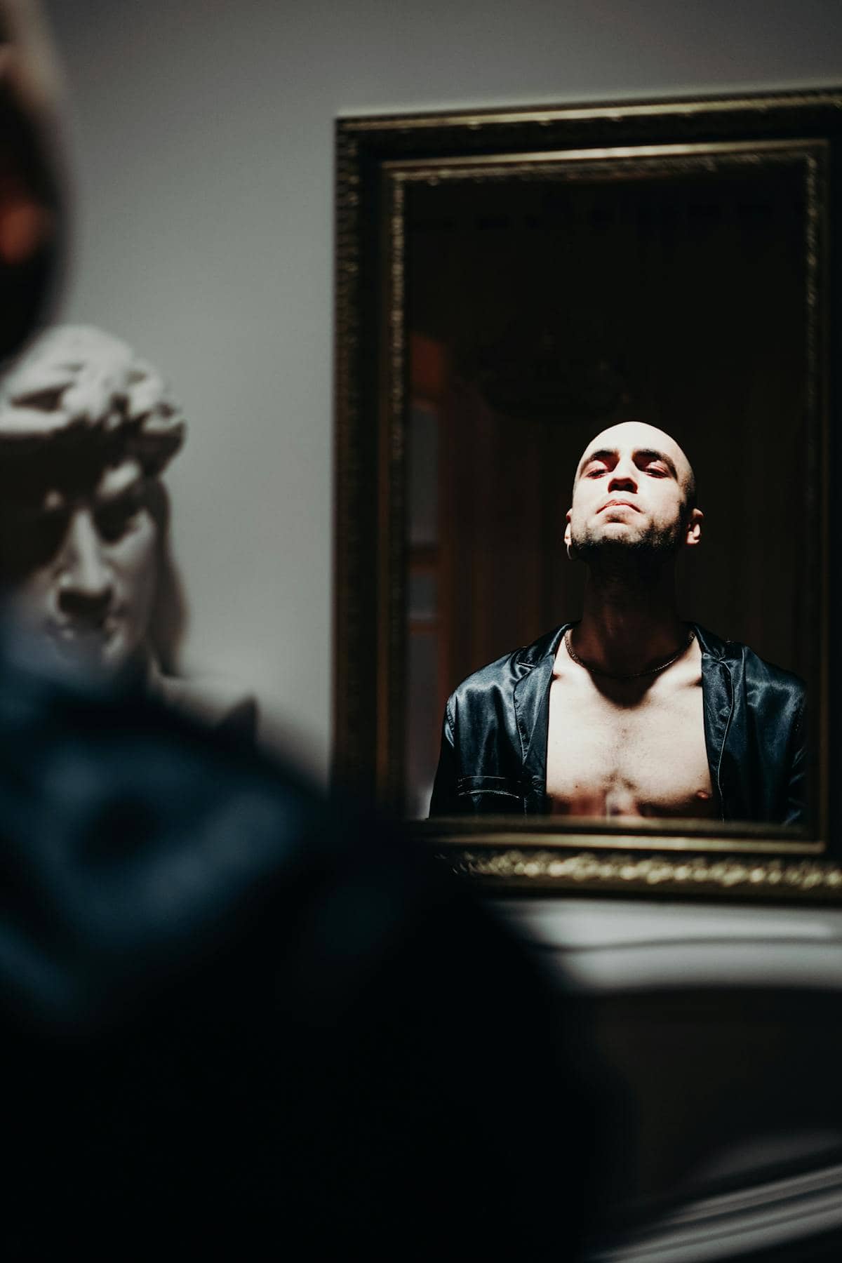 Bald person facing mirror reflection in near-dark room highlighting contemplative introspective atmosphere
