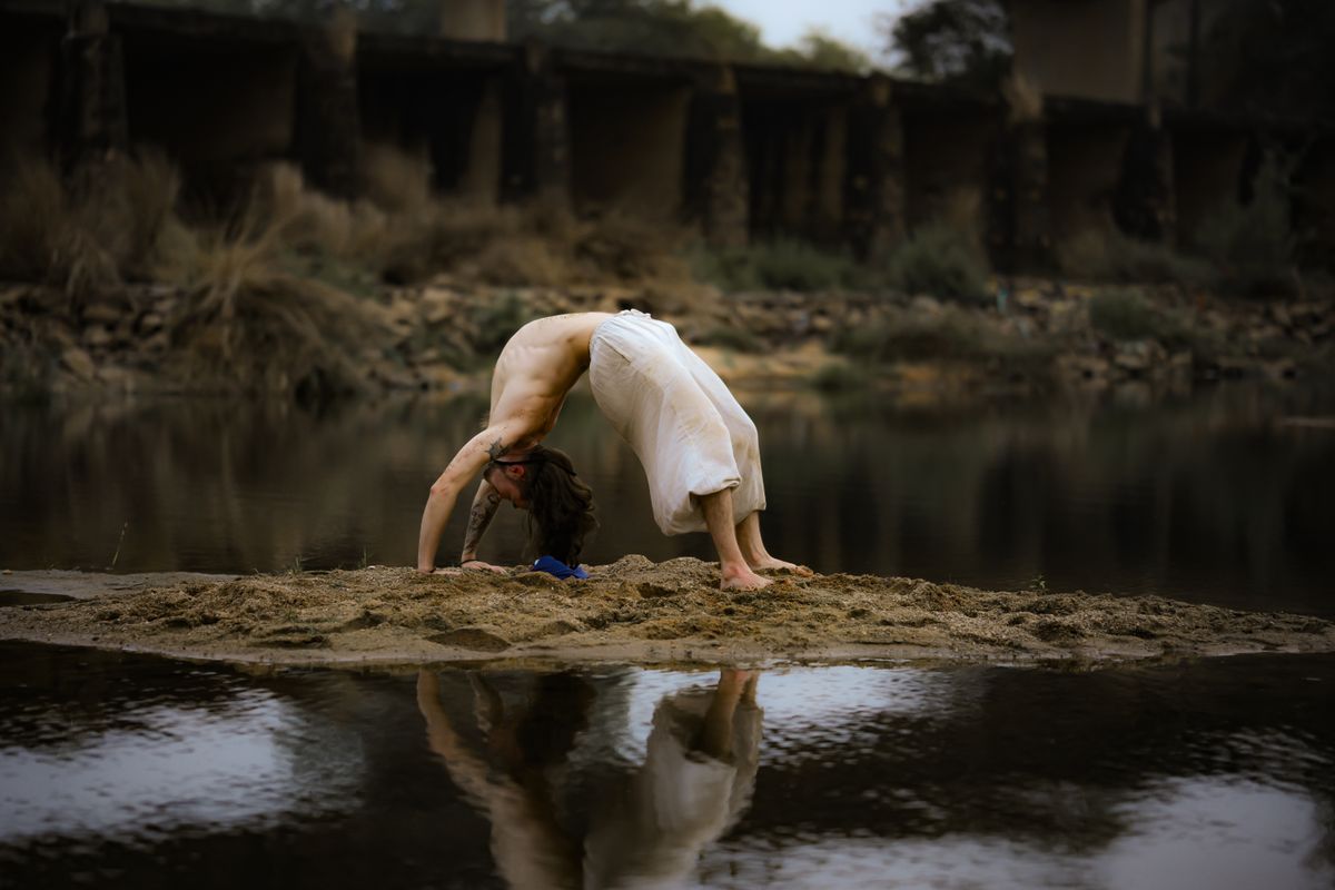A person performs a yoga backbend on a sandy riverbank in a tranquil outdoor setting A person performs a yoga backbend on a sandy riverbank in a tranquil outdoor setting