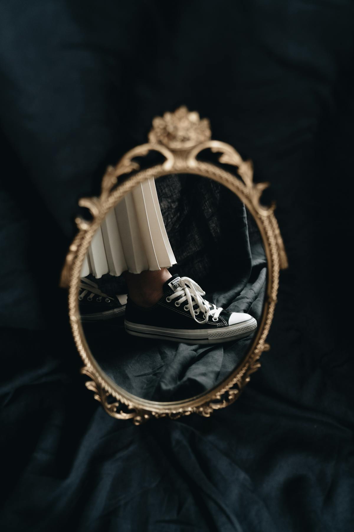Vintage gold-framed mirror reflecting sneakers and skirt on dark fabric artistic composition