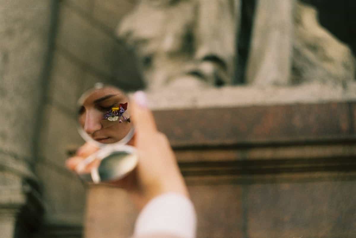 A person's face with flowers reflected in a round mirror held by a hand suggesting spiritual mirror ritual and intentional reflection