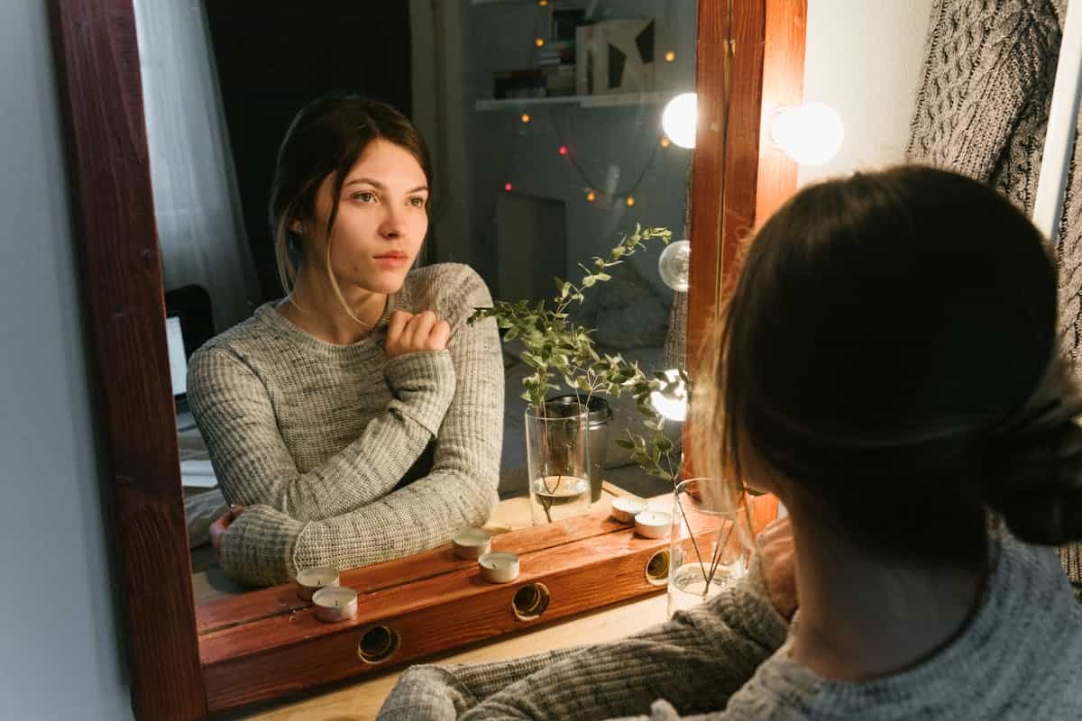 Woman looking at her reflection in warm lighting evoking the self-examination that recurring dreams of mirrors prompt A woman in a cozy sweater contemplates her reflection in a warmly lit vanity mirror evoking the introspective quality of recurring mirror dreams