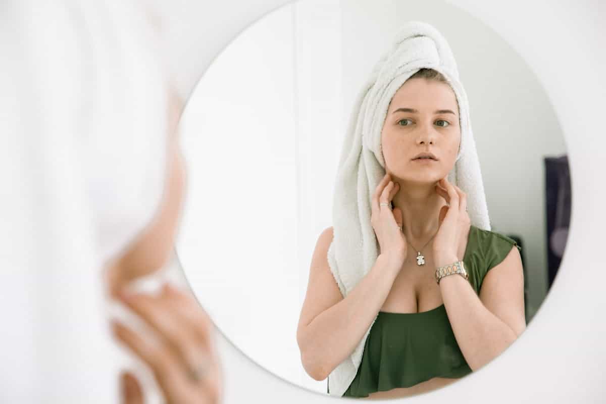 A woman with a white head towel stands at a bathroom mirror in a gentle morning self-care routine, her expression calm and present