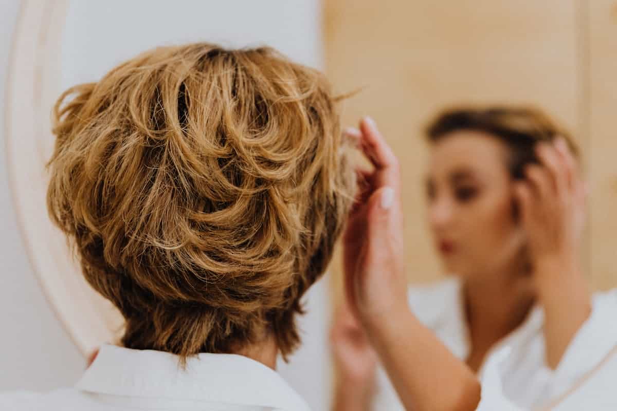 Back view of a woman styling her short hair while looking at her own reflection, representing everyday self-assessment at the mirror