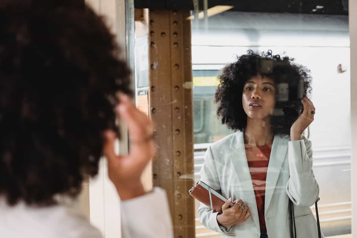 A woman in a white blazer pauses with a thoughtful expression in front of a large mirror, her posture reflective