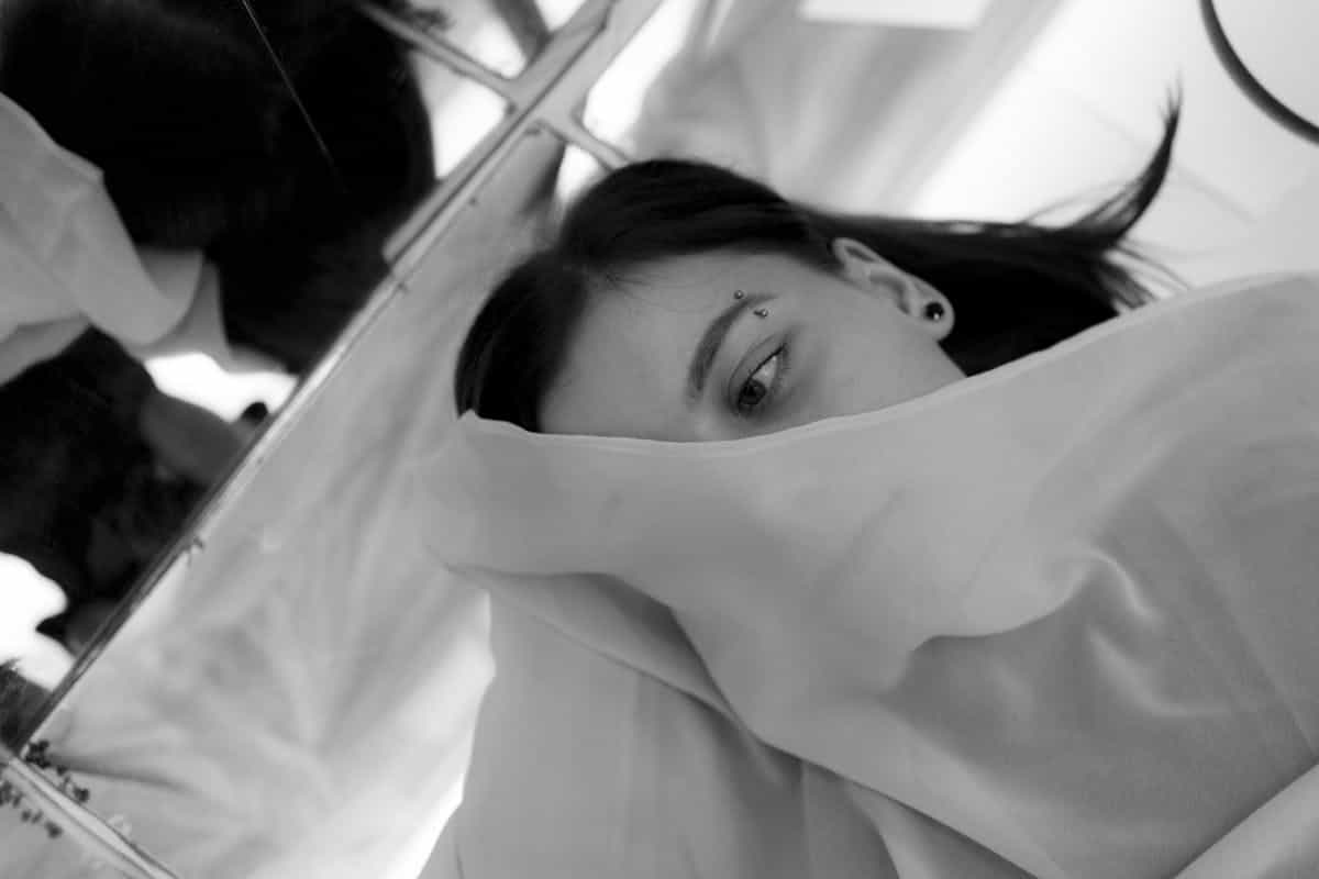 Surreal artistic image of a young woman in white lying under a thin veil on a large mirror representing a dreamlike confrontation with identity