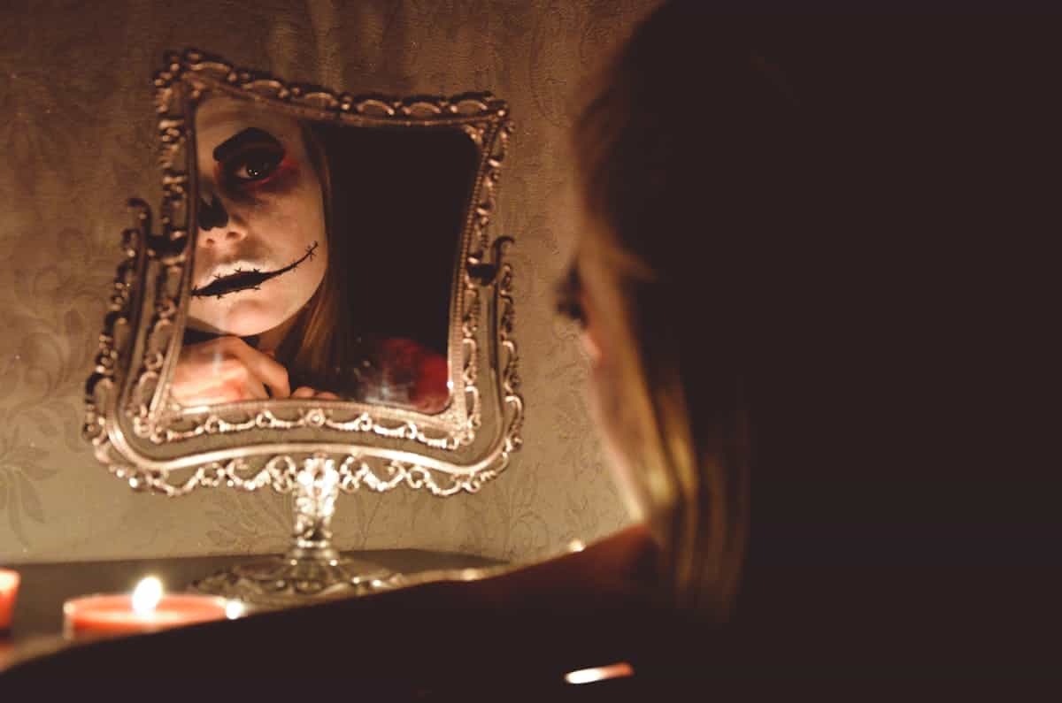 A woman in Halloween makeup stares into an ornate mirror surrounded by candles in a dark room, evoking the uncanny quality of extended mirror gazing