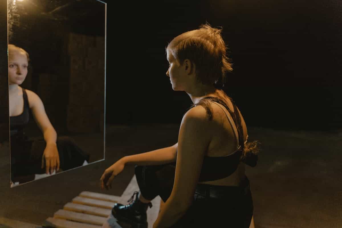 Woman with a shaved head sitting in front of a mirror in a dimly lit room reflecting on her image