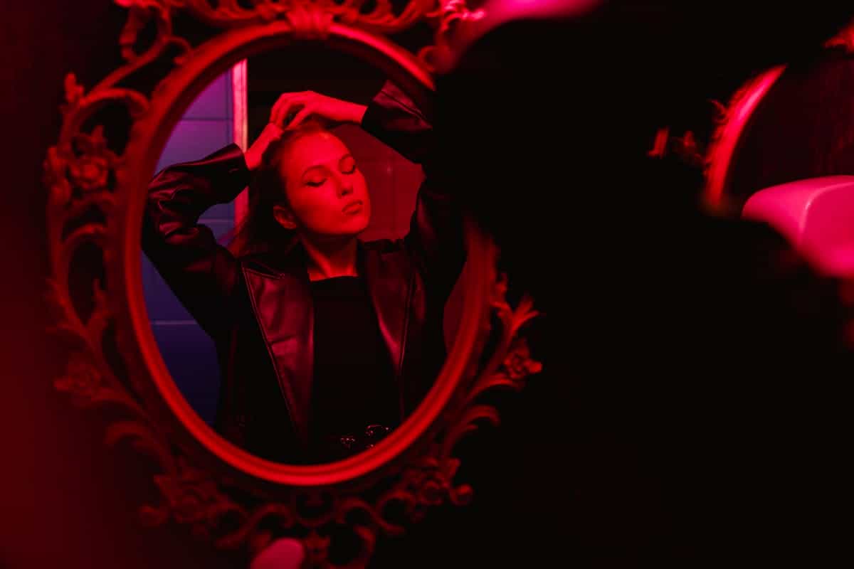 Figure adjusting appearance before moody red-lit mirror illustrating embodied portal metaphors and intentional gaze