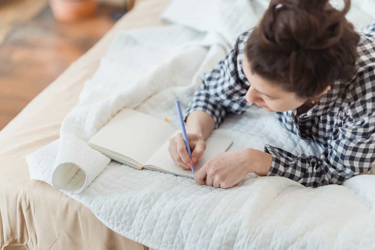 A woman writing in a journal while relaxing on a cozy bed with soft warm lighting evoking the reflective work that follows a cracked mirror dream
