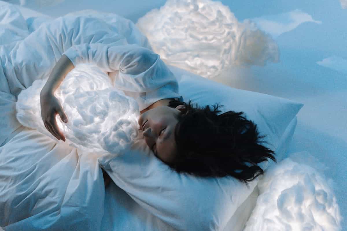 Woman in dreamlike cloud setting evoking the nature of recurring dreams and the sleeping brain returning to unresolved concerns Woman in white holding a glowing cloud pillow in a dreamy pastel blue setting evoking the ethereal quality of sleep and recurring dream states