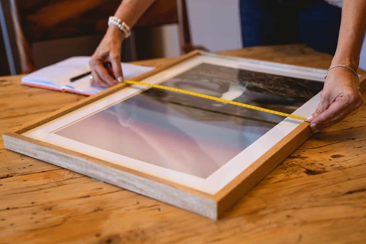 Measuring a picture frame illustrating how to hang magnolia wall art at the right height Hands using a tape measure on a wooden picture frame, illustrating how to measure and hang magnolia wall art