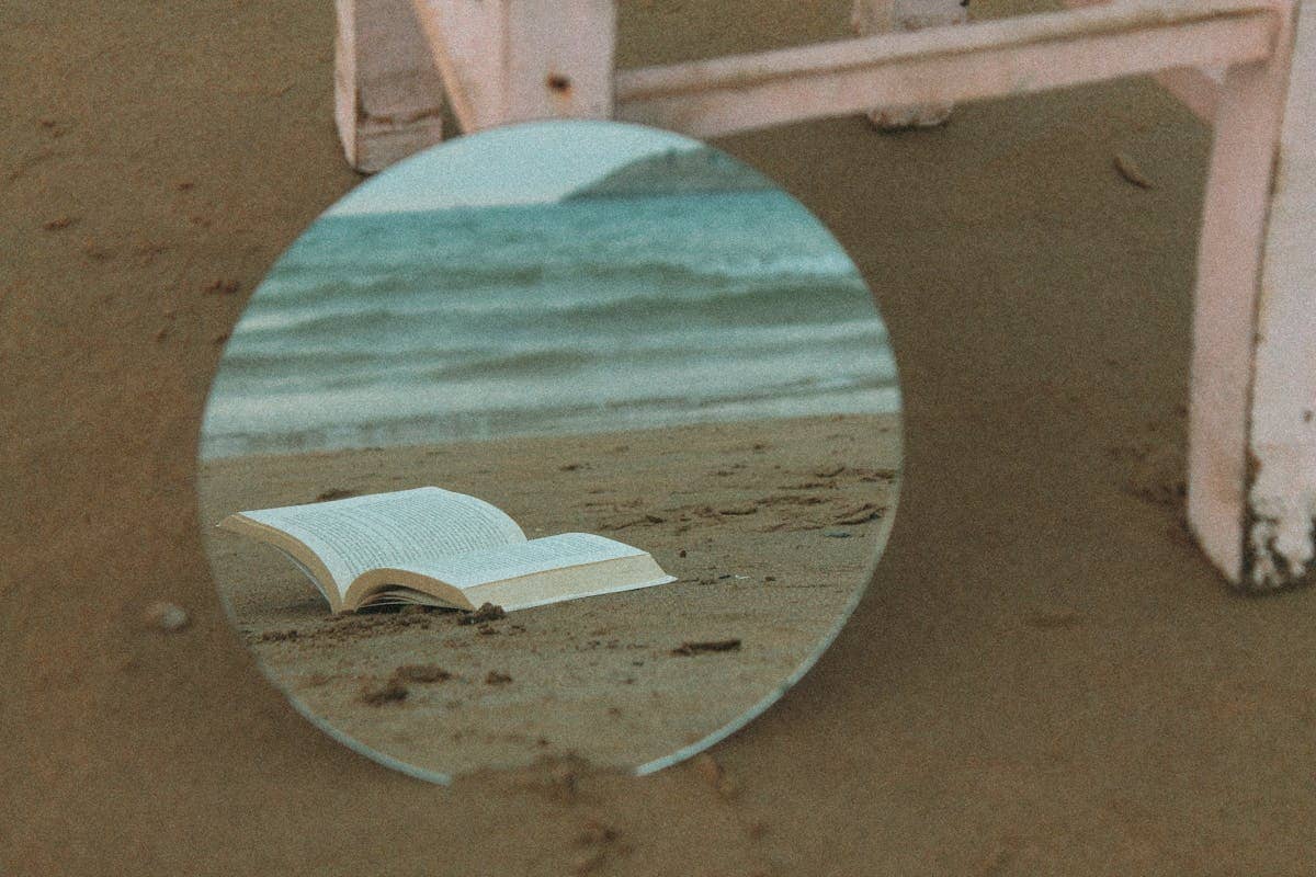 Round mirror reflecting an open book on a sandy beach showing how smooth reflective surfaces create coherent images through the law of reflection