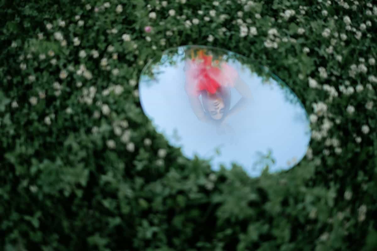 A woman elegantly posed reflected in a round mirror set among lush green foliage in an outdoor setting
