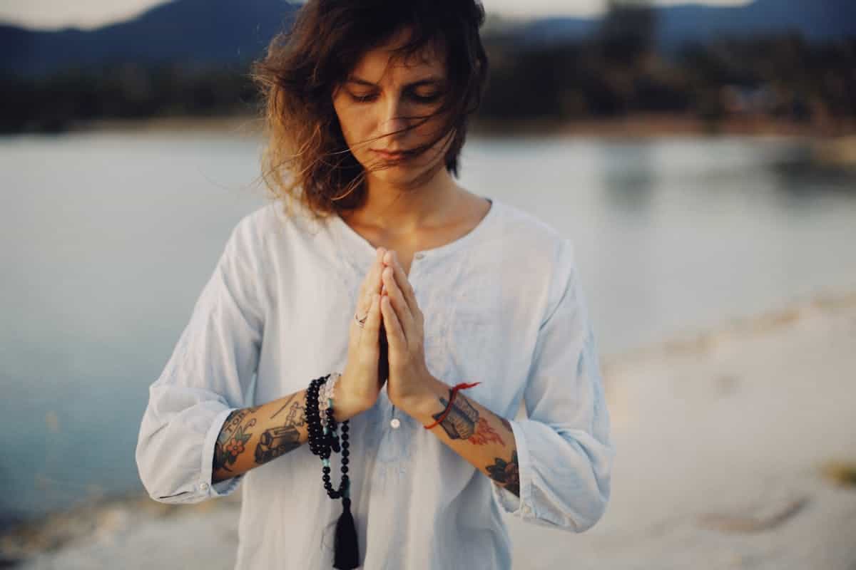 Woman peacefully meditating with prayer beads by a serene lakeside with eyes closed in quiet contemplation, representing the self-reflection practice that a falling mirror spiritually invites