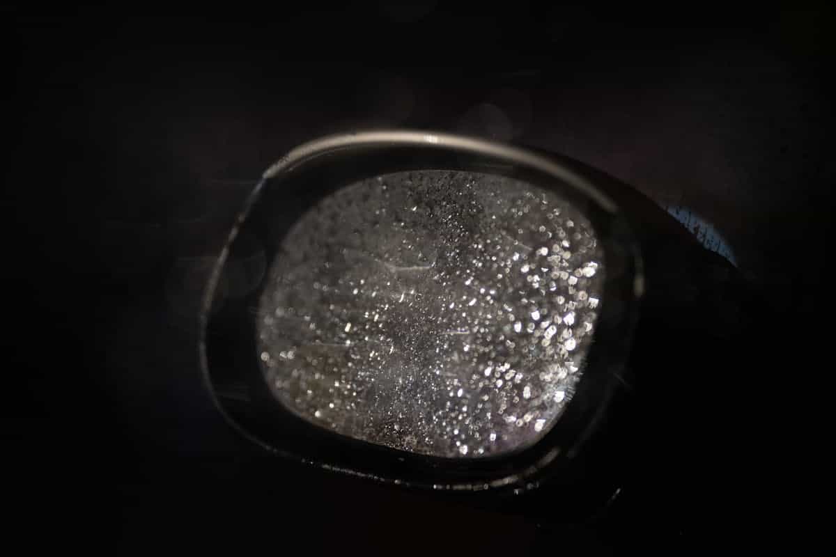 Rain-beaded automotive mirror glass macro emphasizing reflective boundary tension between viewer and wet darkness