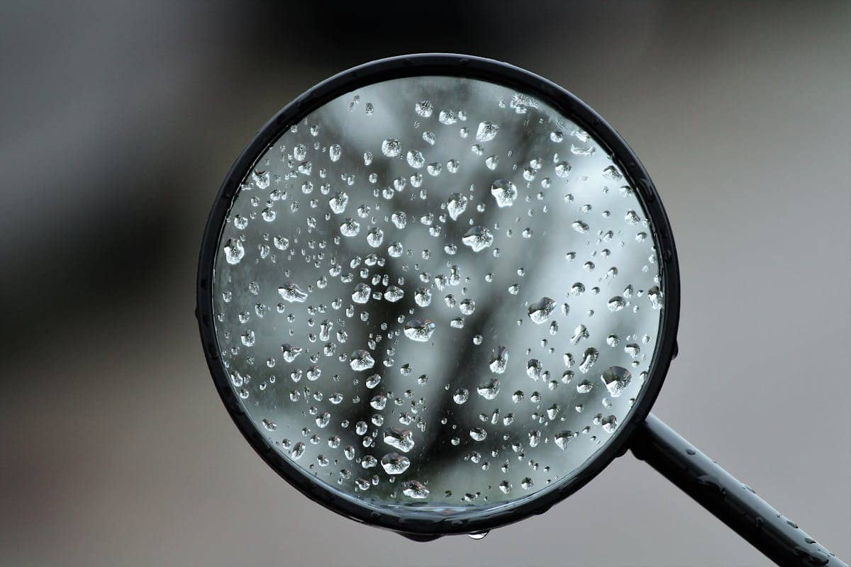 Close-up of dewdrops on a round rearview mirror surface showing the precision reflective coating and how even nanometre-level surface variations affect image coherence and reflectance